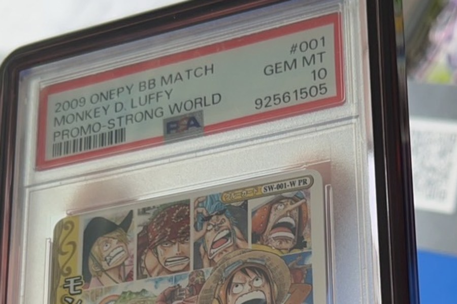 手持一張 PSA 鑑定卡的實拍：標籤寫著 2009 ONEPY BB MATCH、MONKEY D. LUFFY、PROMO-STRONG WORLD、#001、GEM MT 10、流水號 92561505，外層再套一個鯊魚卡磚